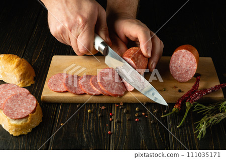 The cook makes sandwiches with salami and bread on the kitchen table. Knife in the hand of a chef for slicing meat sausage The cook makes sandwiches with salami and bread on the kitchen table. Knife in the hand of a chef for slicing meat sausage 111035171