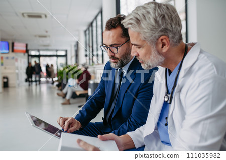 Pharmaceutical sales representative talking with doctor in medical building, presenting new medication on tablet. Hospital director, manager in modern clinic with surgeon. Pharmaceutical sales representative talking with doctor in medical building, presenting new medication on tablet. Hospital director, manager in modern clinic with surgeon. 111035982