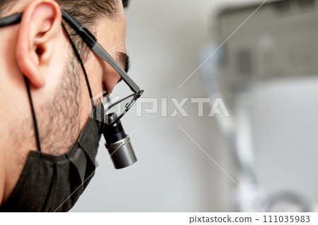 Close-up side portrait of male dentist in surgical mask and dental binocular glasses. Stomatology doctor examines patient's teeth. High-tech dental equipment. Stomatology and dental care concept. Close-up side portrait of male dentist in surgical mask and dental binocular glasses. Stomatology doctor examines patient's teeth. High-tech dental equipment. Stomatology and dental care concept. 111035983