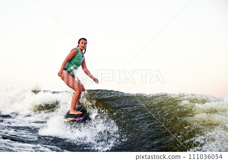 Young beautiful sporty girl in a green life jacket rides a wakeboard on a river or lake near city. Happy athlete girl glides on the wave like a real surfer. Active lifestyle, healthy hobby. 111036054