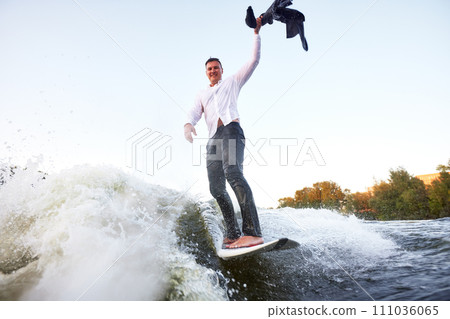 Young man in classic suit rides a wakeboard on the lake near city. Happy clerk waving his jacket escaped from a stuffy office to take up active sport. Best summer leisure after routine work. 111036065