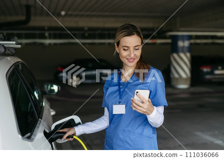 Nurse plugging charger into electric car before going to work. Electric vehicle charging station in front of hospital, modern clinic. Charging at work, workplace. Nurse plugging charger into electric car before going to work. Electric vehicle charging station in front of hospital, modern clinic. Charging at work, workplace. 111036066