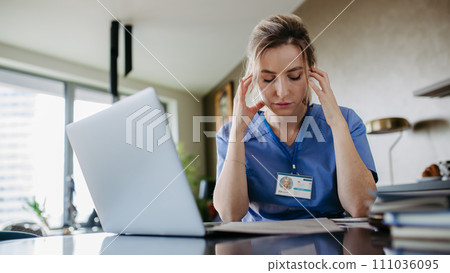 Nurse paying bills online, sitting in living room, working on notebook. Doctor in scrubs dealing with work issues from home, after workday, telemedicine consultation. Work-life balance for healthcare 111036095