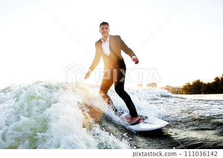 Young man in classic suit rides a wakeboard on the river or lake near city. Clerk escaped from a stuffy office to take up his favorite active sport. Best summer leisure after routine work. Sun flare. 111036142