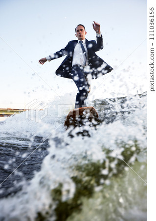 Young man in classic suit rides a wakeboard on the river or lake near city. A careless clerk escaped from a stuffy office to take up his favorite active sport. Best summer leisure after routine work. Young man in classic suit rides a wakeboard on the river or lake near city. A careless clerk escaped from a stuffy office to take up his favorite active sport. Best summer leisure after routine work. 111036156
