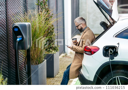 Portrait of businessman waiting while electric car is charging, sitting in car trunk, scrolling on his smartphone, and drinking coffee. An electric vehicle charging station. 111036215