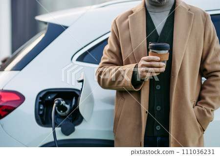 Close up of businessman waiting while electric car is charging, leaning against vehicle, drinking coffee. An electric vehicle charging station. Close up of businessman waiting while electric car is charging, leaning against vehicle, drinking coffee. An electric vehicle charging station. 111036231