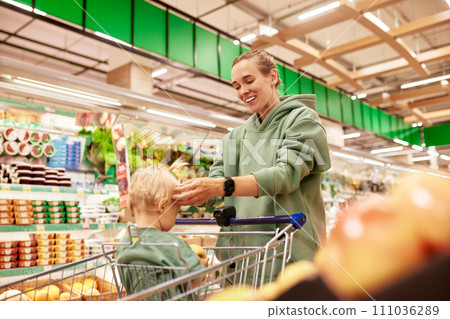 Young attractive mother with her cute baby in shopping trolley walking around supermarket. Beautiful smiling mom and little blonde daughter shopping and having fun. Family weekend, happy childhood. Young attractive mother with her cute baby in shopping trolley walking around supermarket. Beautiful smiling mom and little blonde daughter shopping and having fun. Family weekend, happy childhood. 111036289