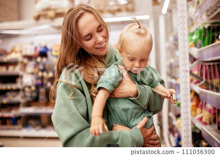 Young mother with cute baby in her hands walks around supermarket. Beautiful mom and little blonde daughter in similar green clothes shopping and having fun. Family weekend, happy childhood. 111036300