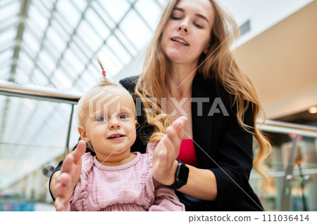 Young pretty mother with a small baby sitting on a bench in a shopping center. Mom and daughter in pink clothes hug, play, relax and have fun while shopping. Family weekend in shopping mall. Young pretty mother with a small baby sitting on a bench in a shopping center. Mom and daughter in pink clothes hug, play, relax and have fun while shopping. Family weekend in shopping mall. 111036414
