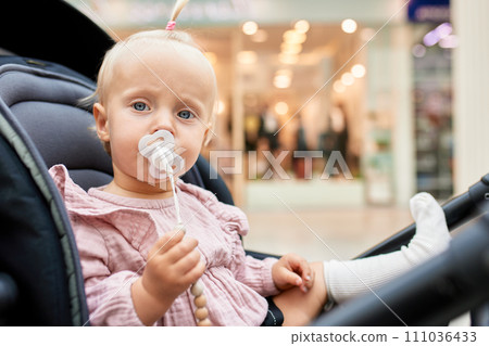 Close-up portrait of a small child in a carriage. Cute little blonde girl in pink clothes with baby pacifier. Mom with a baby in a stroller walking around the mall. Family weekend in shopping mall. Close-up portrait of a small child in a carriage. Cute little blonde girl in pink clothes with baby pacifier. Mom with a baby in a stroller walking around the mall. Family weekend in shopping mall. 111036433
