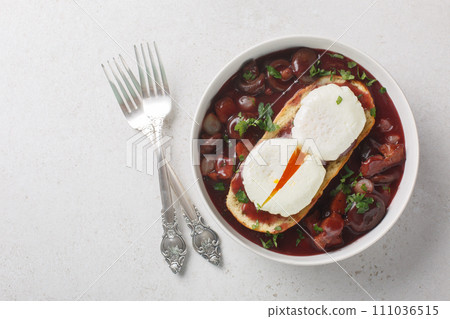 Oeufs en meurette is a classic French dish of poached eggs covered in a rich red wine sauce filled with lardons, mushrooms and onion closeup on the plate. Horizontal top view 111036515