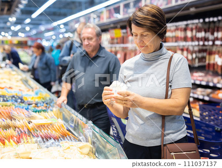 Mature woman diligently choosing fresh cheese in supermarket Mature woman diligently choosing fresh cheese in supermarket 111037137