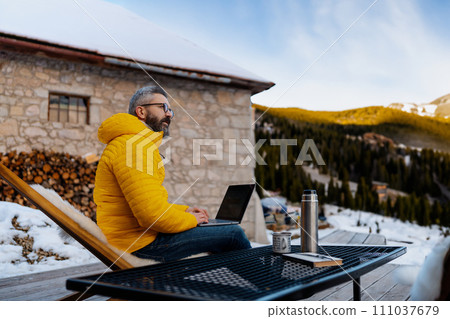 Mature man working from cozy cabin in mountains, sitting on terrace with laptop, enjoying cup of coffee. Concept of remote work from beautiful, peaceful location. Hygge at work. 111037679