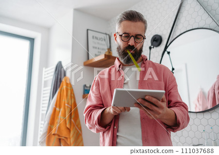 Man shopping online while brushing his teeth in bathroom. Holding tablet, scrolling and buying in eshop, toothbrush in mouth. Man shopping online while brushing his teeth in bathroom. Holding tablet, scrolling and buying in eshop, toothbrush in mouth. 111037688