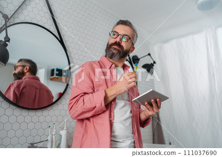 Man shopping online while brushing his teeth in bathroom. Holding tablet, scrolling and buying in eshop, toothbrush in mouth. Man shopping online while brushing his teeth in bathroom. Holding tablet, scrolling and buying in eshop, toothbrush in mouth. 111037690