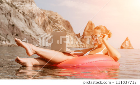 Woman freelancer works on laptop swimming in sea on pink inflatable ring. Happy tourist in sunglasses floating on inflatable donut and working on laptop computer in calm ocean. Remote working anywhere 111037712