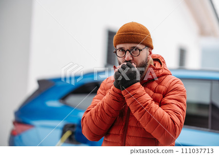 Man charging electric car during cold snowy day. Front view of hansome man feeling cold during winter weather. Charging and driving electric vehicles during winter season. Man charging electric car during cold snowy day. Front view of hansome man feeling cold during winter weather. Charging and driving electric vehicles during winter season. 111037713