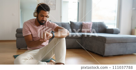 Young man resting after home workout, checking his exercise performance on smartwatch. Morning or evening workout routine, listening to music through wireless headphones. Young man resting after home workout, checking his exercise performance on smartwatch. Morning or evening workout routine, listening to music through wireless headphones. 111037788