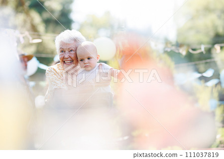 Great-grandmother holding little baby in her arms. Family summer garden party. 111037819