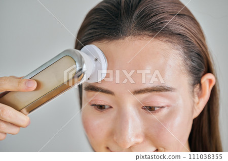 A woman in her 30s with long hair doing skin care using a facial beauty device 111038355