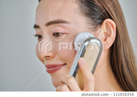 A woman in her 30s with long hair doing skin care using a facial beauty device A woman in her 30s with long hair doing skin care using a facial beauty device 111038358
