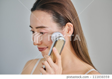 A woman in her 30s with long hair doing skin care using a facial beauty device A woman in her 30s with long hair doing skin care using a facial beauty device 111038359