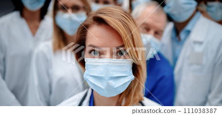 Portrait of beautiful female doctor with surgical mask standing in front of team of doctors. Healthcare team with doctors, nurses, professionals in medical uniforms and face masks in hospital. 111038387