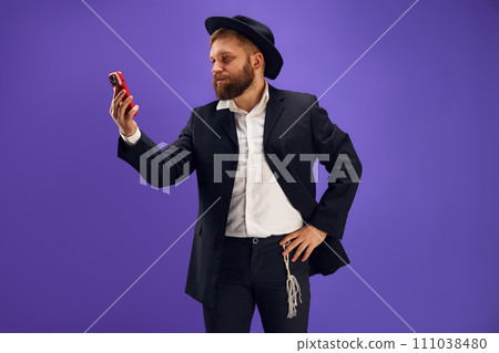 Bearded young Jewish man emotionally looking on mobile phone screen, reading news and messages against purple studio background Bearded young Jewish man emotionally looking on mobile phone screen, reading news and messages against purple studio background 111038480
