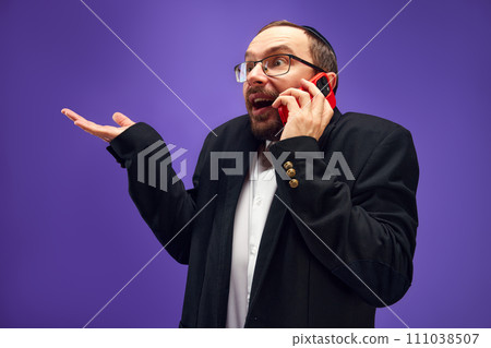 Excited man in glasses and yarmulke emotionally talking on phone, greeting with holiday against purple studio background 111038507