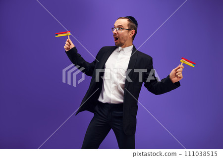 Positive, excited young Jewish man in in glasses standing with noisemaker against purple studio background 111038515