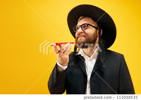 Bearded young man in hat with sidelock recording voice message on phone, greeting with holiday against yellow studio background 111038535