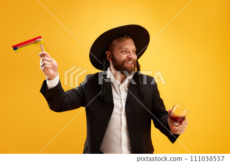 Cheerful young Jewish man in hat, with sidelocks holding noisemaker, against yellow background. Celebration Cheerful young Jewish man in hat, with sidelocks holding noisemaker, against yellow background. Celebration 111038557