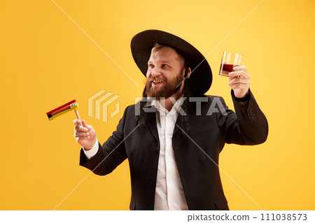 Cheerful young Jewish man in hat, with sidelocks holding noisemaker, against yellow background. Celebration 111038573