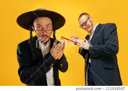Two emotional Jewish man, friends in suits, yarmulke, with noisemaker having fun, celebrating against yellow studio background Two emotional Jewish man, friends in suits, yarmulke, with noisemaker having fun, celebrating against yellow studio background 111038587