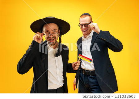 Two emotional Jewish man, friends in suits, yarmulke, with noisemaker having fun, celebrating against yellow studio background 111038605