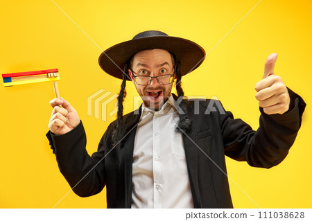 Emotional, excited young Jewish man in hat, with sidelocks, in glasses standing with noisemaker against yellow studio background Emotional, excited young Jewish man in hat, with sidelocks, in glasses standing with noisemaker against yellow studio background 111038628
