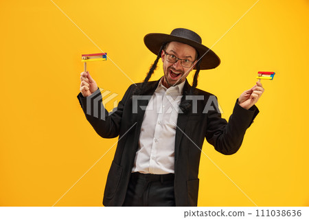 Cheerful Jewish man in hat, with sidelocks holding noisemaker, against yellow background. Celebration Cheerful Jewish man in hat, with sidelocks holding noisemaker, against yellow background. Celebration 111038636