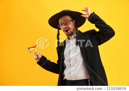 Cheerful Jewish man in hat, with sidelocks holding noisemaker, against yellow background. Celebration 111038639
