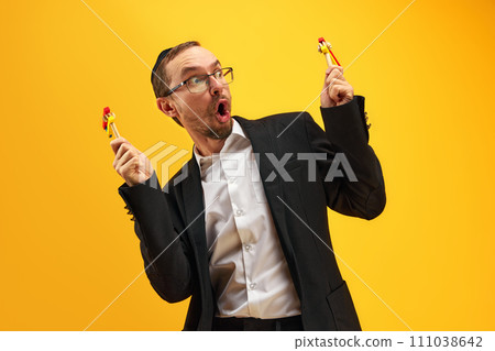 Portrait of man in his 30s wearing glasses, yarmulke and playing with noisemaker, gragger, expressing emotions against yellow studio background 111038642