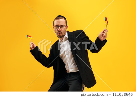 Portrait of man in his 30s wearing glasses, yarmulke and playing with noisemaker, gragger, expressing emotions against yellow studio background 111038646