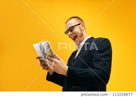 Emotional Jewish young man in glasses, looking on tablet with excitement, reading news against yellow studio background Emotional Jewish young man in glasses, looking on tablet with excitement, reading news against yellow studio background 111038677