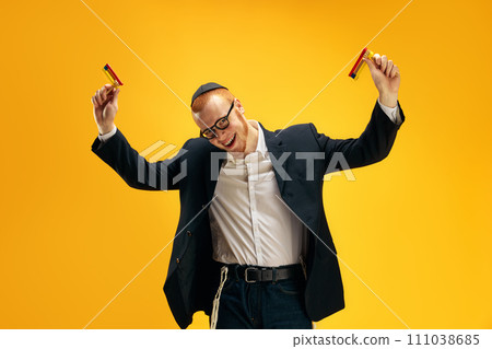 Young redhead Jewish man in glasses, yarmulke playing with wooden noisemaker, gragger against yellow studio background Young redhead Jewish man in glasses, yarmulke playing with wooden noisemaker, gragger against yellow studio background 111038685