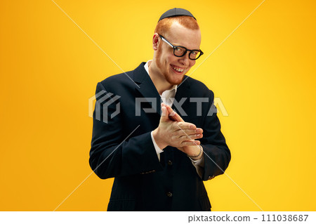 Portrait of happy young redhead Jewish man in glasses and yarmulke cheerfully posing against yellow studio background 111038687