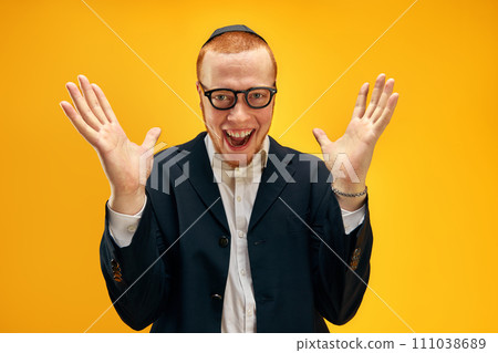 Portrait of happy young redhead Jewish man in glasses and yarmulke cheerfully posing against yellow studio background Portrait of happy young redhead Jewish man in glasses and yarmulke cheerfully posing against yellow studio background 111038689