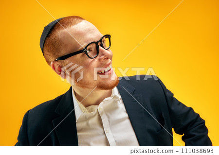 Portrait of smiling, happy young Jewish man in yarmulke, glasses and suit standing against yellow studio background 111038693