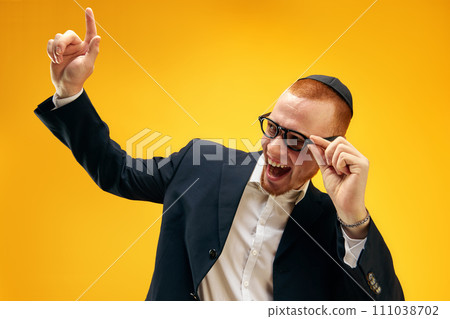 Portrait of smiling, happy young Jewish man in yarmulke, glasses and suit raising finger up against yellow studio background 111038702
