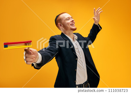 Happy, positive young Jewish man in yarmulke, black suit and with wooden noisemaker celebrating Purim against yellow studio background Happy, positive young Jewish man in yarmulke, black suit and with wooden noisemaker celebrating Purim against yellow studio background 111038723