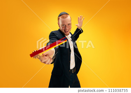 Portrait of emotional young Jewish man in yarmulke, black suit and with wooden noisemaker celebrating Purim against yellow studio background Portrait of emotional young Jewish man in yarmulke, black suit and with wooden noisemaker celebrating Purim against yellow studio background 111038735