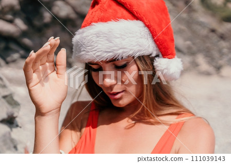 Woman summer travel sea. Happy tourist in red bikini and Santas hat enjoy taking picture outdoors for memories. Woman traveler posing on the beach surrounded by volcanic mountains, sharing travel joy Woman summer travel sea. Happy tourist in red bikini and Santas hat enjoy taking picture outdoors for memories. Woman traveler posing on the beach surrounded by volcanic mountains, sharing travel joy 111039143